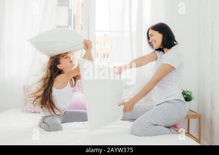 Innen- schuss von angenehmen suchen europäische Weibchen hat Freude zusammen mit ihrer kleinen Tochter, haben, Kissen, Kampf im Schlafzimmer, Pose in der gemütlichen geräumigen hellen Ro Stockfoto