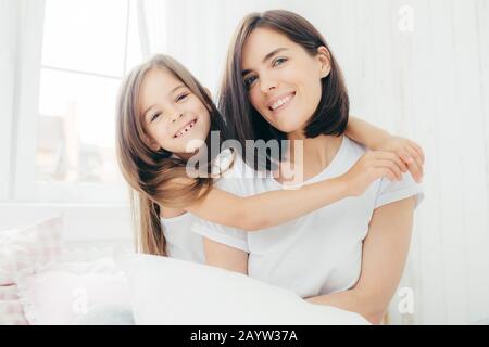 Portrait von Happy weißen Mutter mit charmantem Lächeln und ihrer kleinen Tochter umarmt liebe Mama, sich in guter Stimmung, Haltung auf bequemen Bett agai Stockfoto