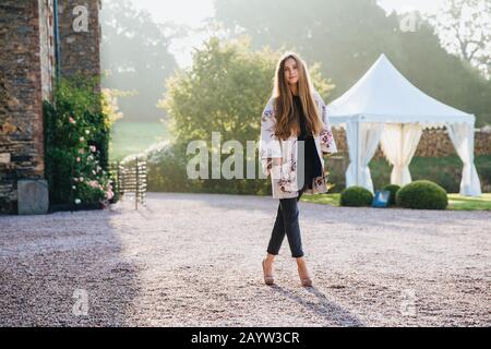 Portrait der angenehm aussehenden schlanken Frau trägt cape und hohe, betuchte Schuhe, steht gekreuzte Beine, stellt sich draußen gegen die alte Gegend, hat Vertrauen Stockfoto