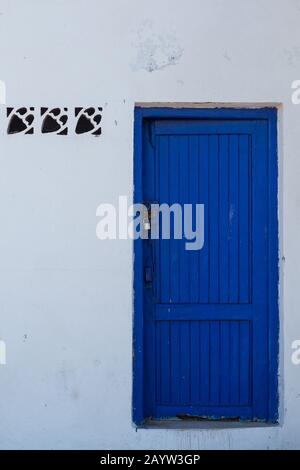 Weiße Fassade eines Hauses mit einer hellblauen Holztür. Drei Fliesen in der Wand neben der Tür. Altstadt - Medina, Essaouira, Marokko. Stockfoto