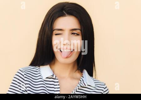 Lustige fröhliche Brunette-Frau zeigt Zunge, blinkt mit Auge, macht Grimace, trägt Hosenträger, hat lange gerade dunkle luxuriöse Haare, beiläufig gekleidet, isol Stockfoto