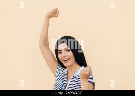 Junge glückliche Frau mit dunkellangem Haar wirft Fäuste mit freudiger Ausdrucksweise auf, hat einen attraktiven Look, trägt lässige Kleidung, freut sich über ihren Erfolg, hat Triump Stockfoto