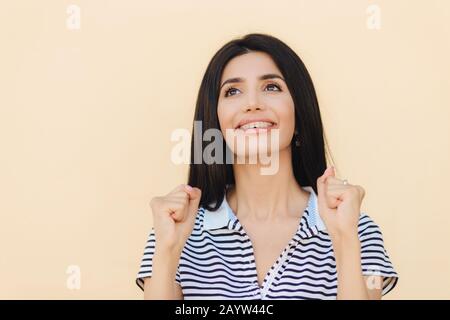 Träumerisch positives Weibchen mit schwarzen geraden Haaren, hält die Hände in Fäusten, blickt glücklich nach oben, glaubt an etwas, isoliert über beigefarbenem Hintergrund. B. Stockfoto