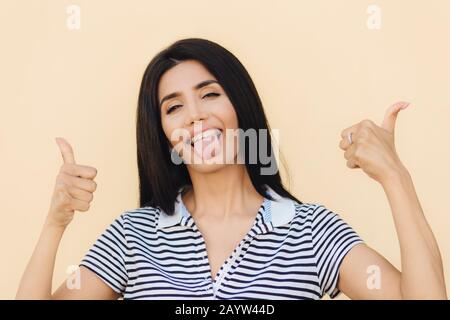 Fröhliche lustige junge Frau mit angenehmem Aussehen, Gesten mit beiden Händen, zeigt gutes Zeichen und Zunge, blinkt mit Auge, hat lustigen Look, isolierte Ove Stockfoto