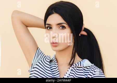 Hübsches Weibchen mit dunklem Haar, Make-up, Puts Lippen, macht Pony Schwanz, hat eine gesunde Haut, gekleidet in lässiger Kleidung, isoliert über hellem Hintergrund. Peopl Stockfoto