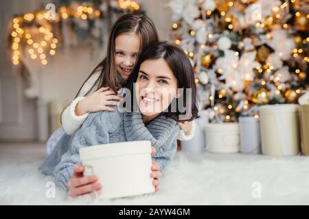 Dieses Geschenk ist für Dich! Happy kleines Kind umarmt ihre liebevolle Mutter, die hält gewickelt vorhanden, steht gegen gestalteten Hintergrund mit Girlanden Stockfoto