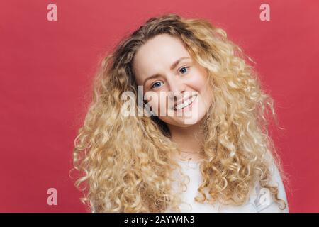 Glücklich erfreute gute Loking-Frau mit positivem Ausdruck, lockig buschigen Haaren, lächelt freudig an der Kamera, isoliert über dunkelrosa Hintergrund. Aktiv Stockfoto
