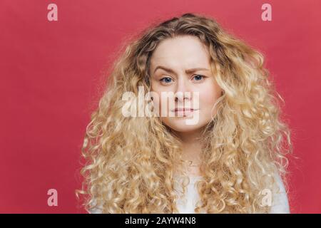 Junge attraktive Frau hat unzufriedene Ausdrucksweise, hebt Augenbraue in Verwilderung, ist unzufrieden mit etwas, hat buschige lockige Haare, isoliert OV Stockfoto