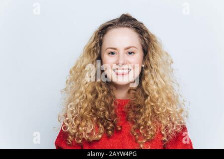 Innenschuss von fröhlicher, lockig blonder Frau mit angenehmem Lächeln und fröhlichem Ausdruck, bekleidet mit warmrotem Pullover, Modelle vor weißem Hintergrund. Peo Stockfoto