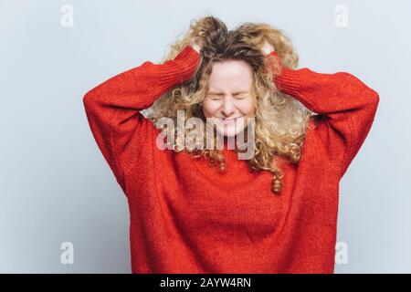 Stressige frustrierte blonde Frau reißt Haare aus, bedauert Fehlstacheln, drückt negative Emotionen aus, trägt einen lockeren roten Pullover, isoliert über weiße Studien Stockfoto