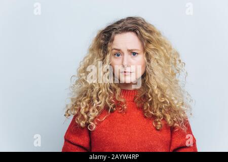 Foto einer unzufriedenen, unglücklichen jungen Frau mit lockig buschigen blonden Haaren, Stirnrunzeln in verwirrender Weise, trägt legere Kleidung, Models gegen weißes Studio Stockfoto