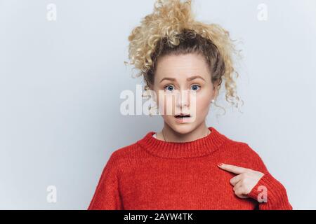 Schockiert verblüffte junge Frau mit überraschtem Ausdruck, deutet auf einen leeren roten Pullover hin, wirbt neues Outfit, posiert vor weißem Studiohintergrund Wi Stockfoto