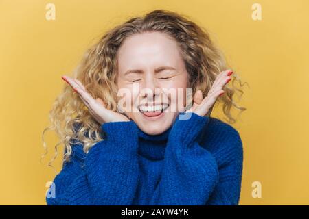 Fröhliche, lockige Frau mit gesunder Haut, lacht laut, hält die Augen geschlossen, ist in hohem Geist, hört lustige Witze von Gesprächspartner, trägt blaue swe Stockfoto