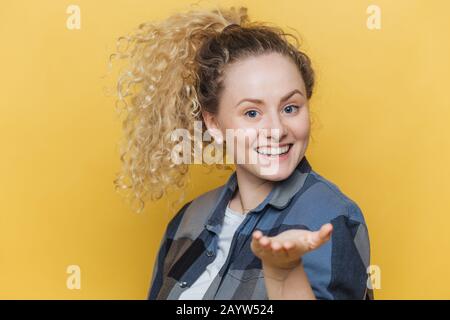 Schöne, freudige Frau streckt sich die Hand im Vordergrund, hat ein breites Lächeln, zeigt perfekte weiße Zähne, trägt ein kariertes Hemd, posiert gegen den gelben Backgroschen Stockfoto