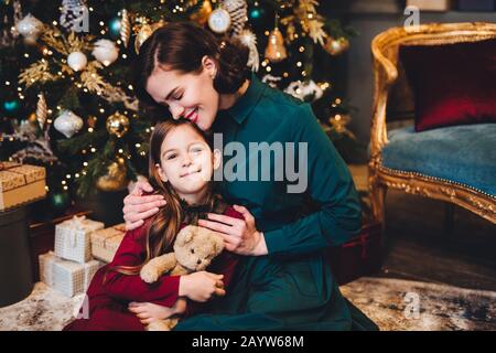 Liebevolle Mutter umarmt ihre kleine Tochter zusammen in der Nähe von geschmückten Weihnachtsbaum sitzen. Kleine niedliche Mädchen, froh, dass das neue Jahr bereits erhalten Stockfoto