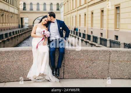 Liebevolles Ehepaar sitzt auf Brückenschlag genießen frische Luft, haben gute Beziehung, küssen sich. Junge Frau in weißem Hochzeitskleid Reci Stockfoto