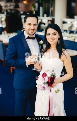 Brautpaar mit Gläsern Champagner, feiern ihre Hochzeit, warten auf Gäste in Festsaal, lächeln angenehm auf die Gesichter. Putsch verheiratet Stockfoto