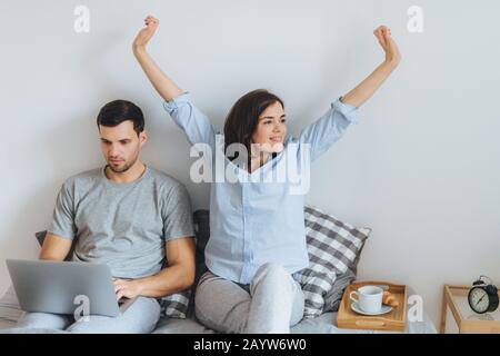 Junges Ehepaar Morgen im Bett verbringen: schöne Brünette Frauen erstreckt sich und ihr Mann mit ernsten Ausdruck verwendet Laptops für die Fernbedienung Stockfoto