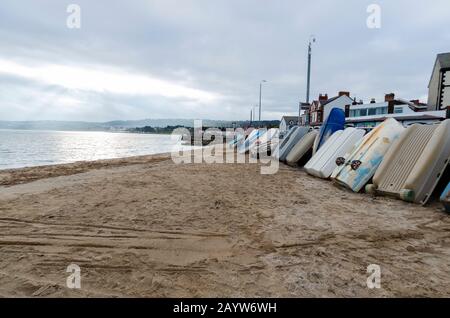 Kleine Boote, die am Strand von Rhos am Meer gegen die Meereswand stehen. Die Sonne versucht durch den grauen Winterhimmel zu brechen. Stockfoto