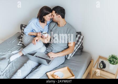 Schönes romantisches Paar gehen zu küssen, an jedem anderen aussehen, wie auf dem Bett sitzen, mit Laptop Computer für die Wiedergabe von Filmen, Kaffee trinken, Zweisamkeit geniessen und r Stockfoto