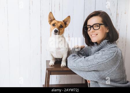 Foto von adorable froh, zufrieden Weibliche trägt eine Brille und Lose warmen Pullover, steht in der Nähe von ihren Liebling Hund, auf Stuhl sitzt, Blick in die Kamera, Goin Stockfoto
