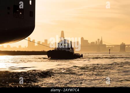 Schleppboot und Containerschiff ziehen gegen die untergehende Sonne und die Skyline von San Francisco in den Hafen von Oakland. Stockfoto