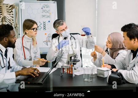 Das Team von multiethnischen Wissenschaftlern in Labormänteln in modernen Labors führt medizinische Forschung auf der Suche nach Impfstoff oder neuem Medikament durch. Reifer Mann im Maskenschauen Stockfoto