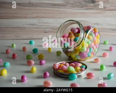 Ein Glas Süßigkeiten-Glas, gefüllt mit gesprenkelten Pastellgelstinbohnen, die zu Ostern auf eine weiße Oberfläche mit Holzhintergrund auslaufen. Stockfoto