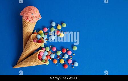 Festlicher bunter Partylebensmittelhintergrund mit Kopierraum auf Blau mit Erdbeereis in einem Waferkegel und regenbogenfarbenen, zuckerbeschichteten Süßigkeiten spil Stockfoto