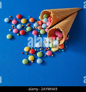 Regenbogenfarbene, zuckerbeschichtete Schokoladenbonbons und bestreuen Spilling aus zwei Wafer-Cornets oder Eiskegel auf einen blauen Hintergrund im quadratischen Format Stockfoto