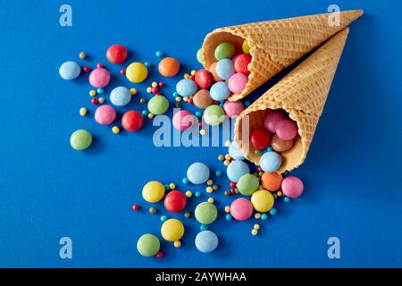 Partyhintergrund mit farbenfroher, zuckerbeschichteter Schokoladenbonbons, die über Blau- und Wafer-Eiskegel verstreut sind, in flacher Laienlebensdauer mit Kopierraum Stockfoto