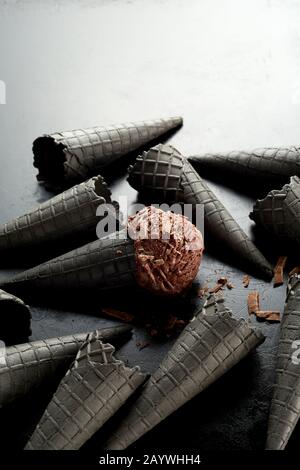 Willkürliche Streuung von grauen Eiskegel mit einer Schokoladenschaufel, die im mittleren Kornett über einem gradienten grauen Hintergrund mit Kopierraum in Flocken bedeckt ist Stockfoto