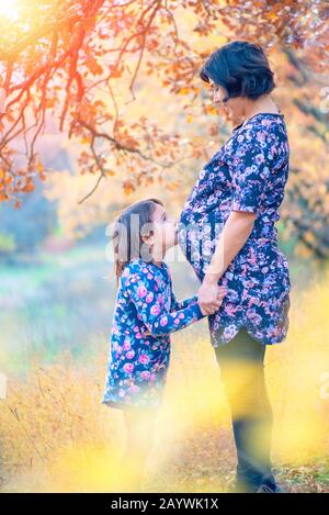 Glückliche Familie. Schwangere Mutter und kleine Tochter umarmen und küssen bei Sonnenuntergang im Herbst im Wald. Gelächter, Glück und Spaß zusammen Stockfoto