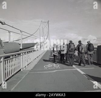 1980er Jahre, eine Gruppe von erwachsenen Wanderern mit vollem Wandergeschirr, die im Straßenbelag und Radabschnitt der Forth Road Bridge, Stirling, Schottland, eine der bedeutendsten Hängebrücken der Welt mit langer Spannweite entlang spazieren. Stockfoto