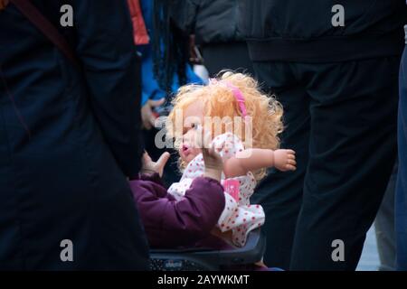 Ein Kind im Kinderwagen wird mit seiner Puppe fotografiert. Es gibt viele Menschen in der Umgebung. Stockfoto