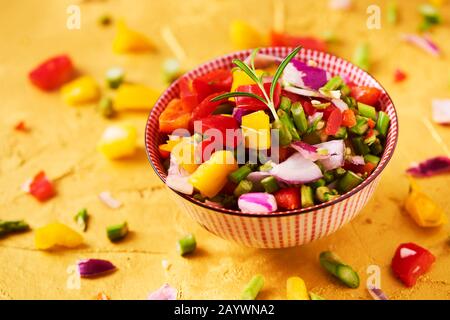 Nahaufnahme einer Schüssel mit einer Mischung verschiedener roher gehackter Gemüse, wie Spargel, Zwiebel oder gelber und roter Paprika, auf einem goldfarbenen, strukturierten Surfac Stockfoto