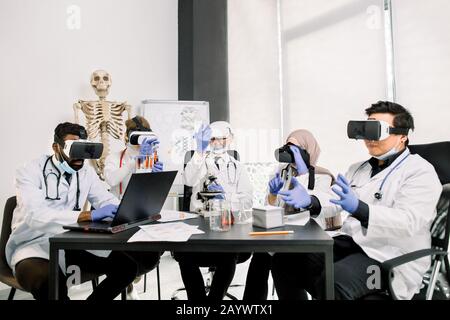 Wissenschaftler mit vr-Brille für Biochemie-Tests im modernen Labor. Afrikanischer Mann arbeitet auf Laptop, kaukasische Frau hält Röhrchen, kaukasischer Mann betrachtet Stockfoto