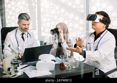 Zwei Medizinstudenten oder junge Wissenschaftler tragen eine VR-Brille, studieren Biochemistie oder Pharmakologie, und ihr männlicher Lehrer betrachtet sie und macht sie Stockfoto