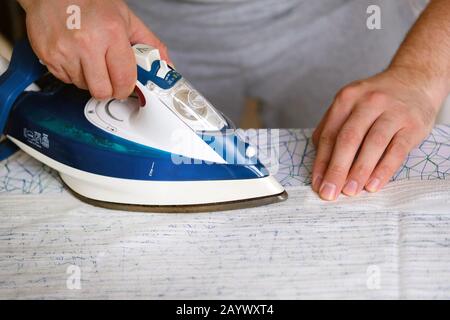 Hausarbeit und Haushaltskonzept. Mann bügelt zu Hause Textil auf dem Bügelbrett. Blaues Eisen in der Hand des Menschen. Stockfoto