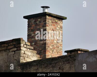 Alter brauner, strukturierter Kamin aus Backstein mit Betondeckelstein und Aluminiumstapel. Beschädigte Ziegelwand. Hellblauer Himmel. Bauliches Wartungskonzept. Stockfoto