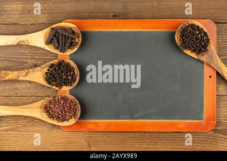 Vier Kochlöffel aus Holz aus olivem Holz mit verschiedenen Pfefferkörnern und einer alten schwarzen Schiefertafel mit Kopierraum auf rustikalem Holzhintergrund Stockfoto