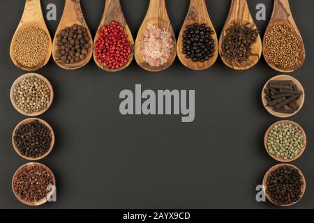 Sieben Kochlöffel aus olivem Holz und sechs kleine Holzschüsseln mit verschiedenen Gewürzen auf schwarzem Hintergrund mit Kopierraum Stockfoto