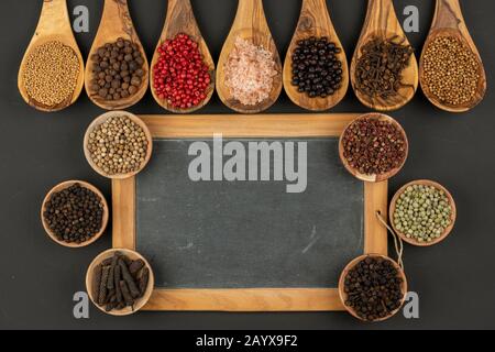 Sieben Kochlöffel aus olivem Holz und sechs kleine Holzschüsseln mit verschiedenen Gewürzen und einer alten schwarzen Schiefertafel mit Kopierraum auf einem Schwarz Stockfoto