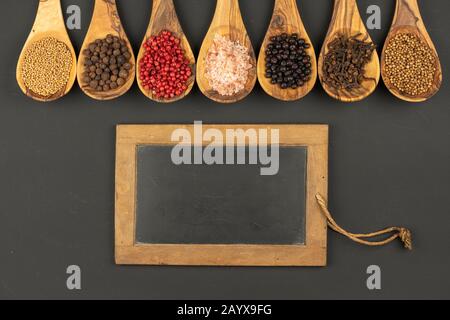 Sieben Kochlöffel aus olivem Holz mit verschiedenen Gewürzen liegen in einer Reihe und eine alte schwarze Schiefertafel mit Kopierraum auf schwarzem Hintergrund Stockfoto