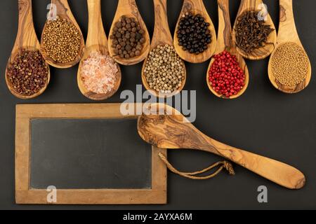 Gewürz und Essen - Neun Kochlöffel aus olivem Holz in einer Reihe mit Gewürz- und Pfeffersorten und einem alten schwarzen Schiefertafel mit Kopierraum Stockfoto