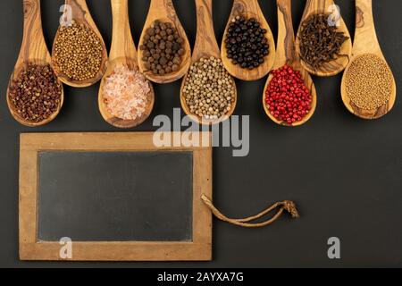 Gewürz und Essen - Neun Kochlöffel aus olivem Holz in einer Reihe mit Gewürz- und Pfeffersorten und einem alten schwarzen Schiefertafel mit Kopierraum Stockfoto