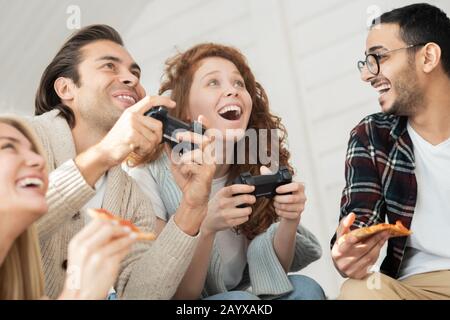 Unten sehen Sie aufgeregt junge Mann und Frau, die Videospiele spielen, während ihre Freunde Pizza essen und für sie rooten Stockfoto
