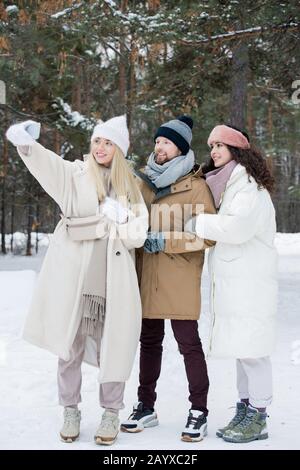 Vertikaler Schuss von jungen Männern und zwei Frauen verbringen zusammen Zeit im Freien am Wintertag, um selfie auf dem Smartphone zu fotografieren Stockfoto