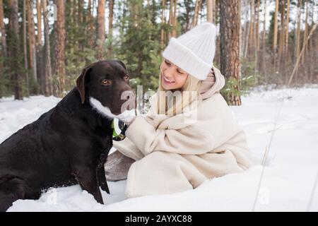 Horizontales Porträt einer fröhlichen Frau mit langen blonden Haaren, die im Schnee sitzen und sich am Wintertag mit ihrem Labrador-Hund lustig machen Stockfoto