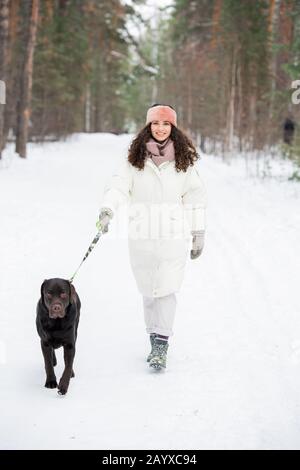 Vertikaler Vollschuss von stilvoller junger Frau, die am Wintertag ihren Labrador Retriever entlang der städtischen Waldstraße spazieren ließ Stockfoto